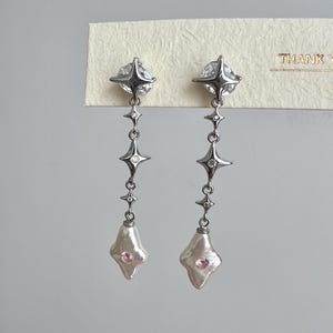 May include: A pair of silver earrings with a dangling star design and a pink pearl at the bottom. The earrings are on a white card with the text "THANK YOU" in black.