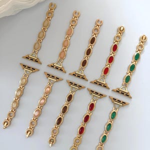 May include: Gold-toned watch bands with oval-shaped gemstones in various colors, including beige, pink, brown, red, and green. The bands have a chain-like design and are displayed on a white surface.