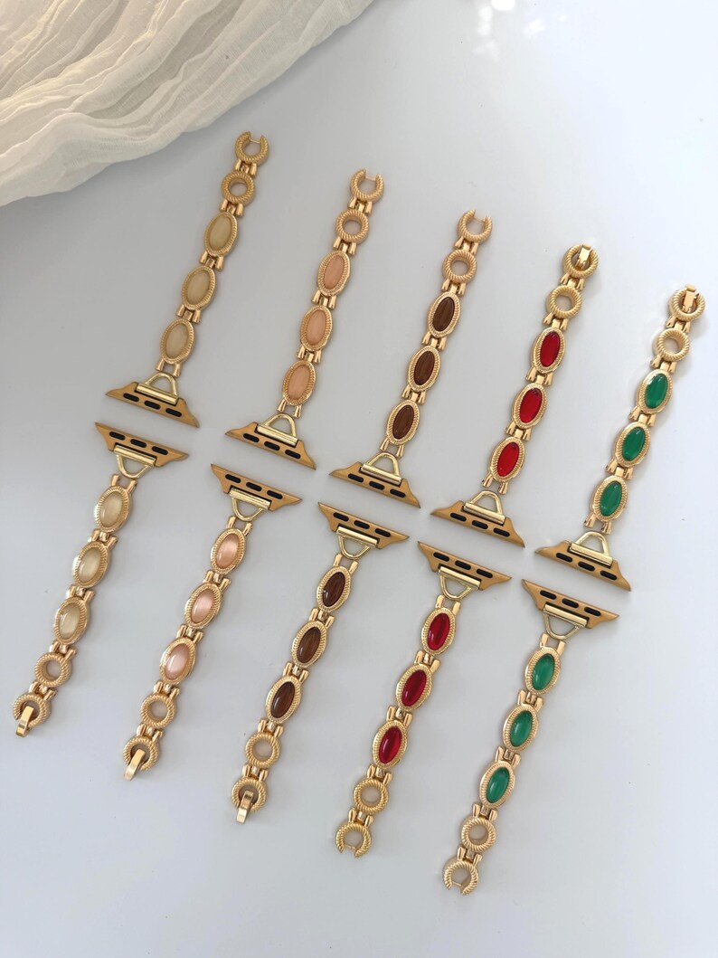 May include: Gold-toned watch bands with oval-shaped gemstones in various colors, including beige, pink, brown, red, and green. The bands have a chain-like design and are displayed on a white surface.