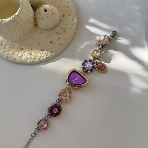 May include: A decorative wristwatch with a purple face and gold-tone bezel. The watch band features various decorative charms, including flowers and gemstones in shades of purple, pink, and gold. The watch is displayed on a white surface.