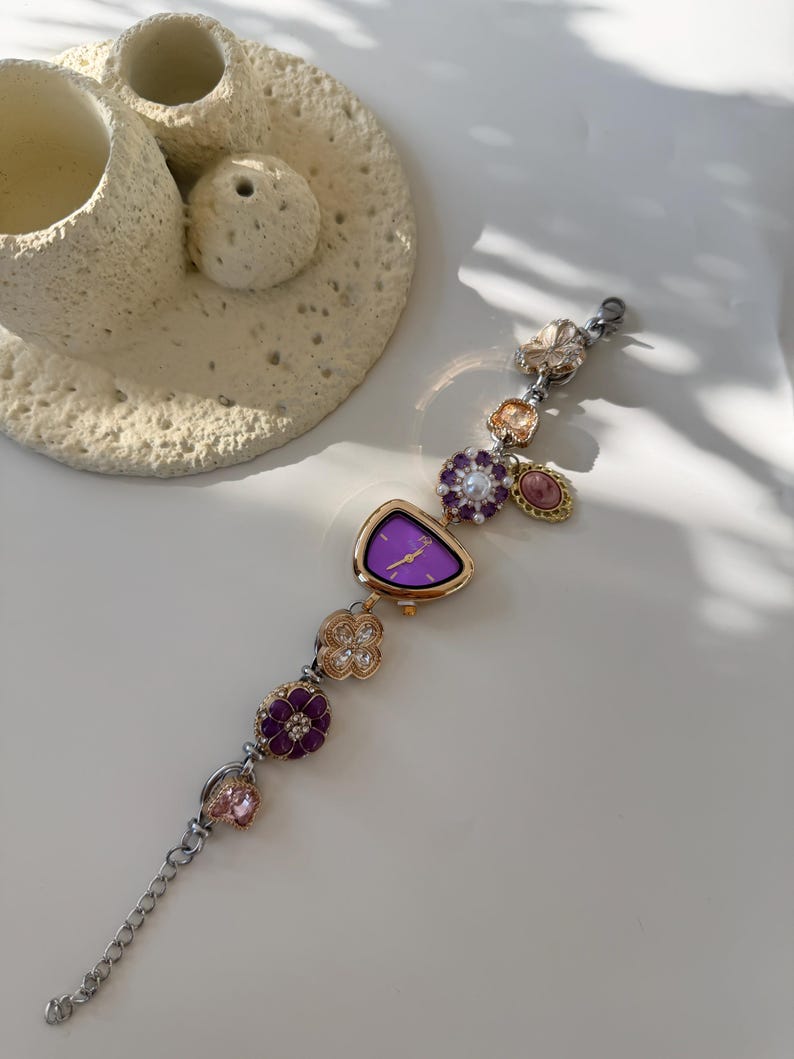 May include: A decorative wristwatch with a purple face and gold-tone bezel. The watch band features various decorative charms, including flowers and gemstones in shades of purple, pink, and gold. The watch is displayed on a white surface.
