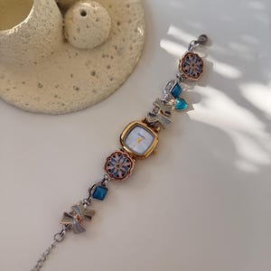 May include: A gold-coloured watch with a square face and a silver band. The band features decorative elements including blue gems, floral designs, and bow-shaped charms. The watch face has the word "LEAFS" printed on it.