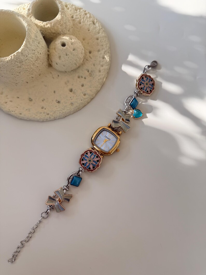 May include: A gold-coloured watch with a square face and a silver band. The band features decorative elements including blue gems, floral designs, and bow-shaped charms. The watch face has the word "LEAFS" printed on it.