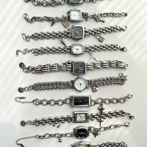 May include: A collection of ten silver-toned wristwatches arranged in rows. Each watch features a unique metal link bracelet, some adorned with small charms like keys, locks, crosses, or stars. The watch faces vary in shape and dial color.