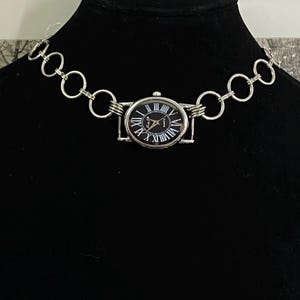 May include: A silver chain necklace with a silver oval watch attached. The watch face is black with white Roman numeral hour markers and a silver second hand.