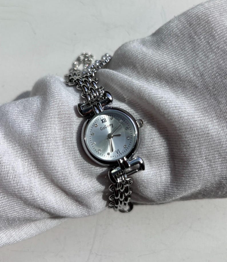 May include: A silver-toned wristwatch with a chain-link band and a round face. The watch face is silver with Roman numeral hour markers and the brand name "GoBoey". The watch is displayed on a white fabric surface.