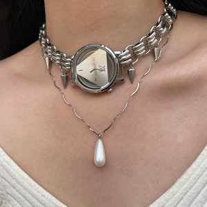 May include: A silver choker necklace with a watch and spikes, layered over a silver chain necklace with a pearl pendant.