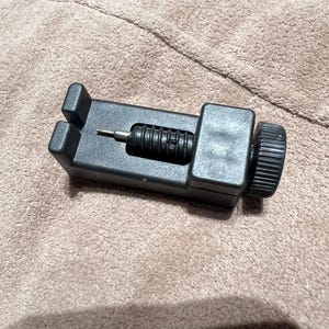 May include: A black watch band link remover tool. The tool has a rectangular base with a screw and a knurled knob on the side. The tool is designed to remove pins from watch bands for resizing or repair.
