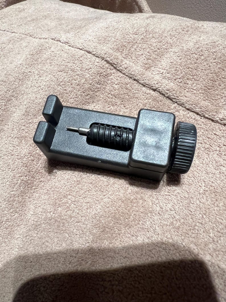 May include: A black watch band link remover tool. The tool has a rectangular base with a screw and a knurled knob on the side. The tool is designed to remove pins from watch bands for resizing or repair.
