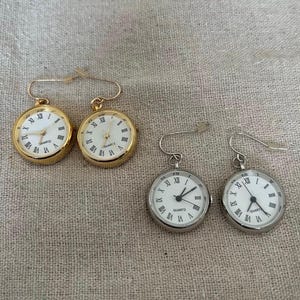 Könnte beinhalten: Ein Paar Ohrringe aus Gold und Silber mit Zifferblättern. Die goldenen Ohrringe haben römische Ziffern und die silbernen Ohrringe arabische Ziffern. Beide Ohrringe zeigen "Quartz" auf dem Zifferblatt.