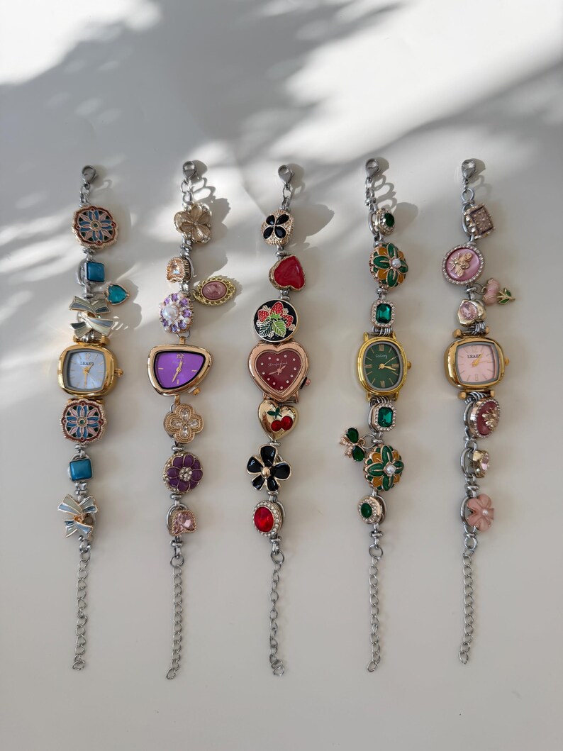 May include: Five decorative charm bracelets with watch faces. Each bracelet has a silver chain and various colourful charms, including flowers, hearts, and gemstones. The watch faces have different shapes and colours, with gold-tone metal cases.