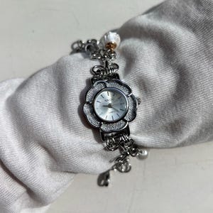 May include: A silver-toned quartz watch with a flower-shaped face and a linked bracelet. The watch face is white with black numerals and hands. The brand name "NAIDU" is printed on the watch face. The bracelet has decorative charms.