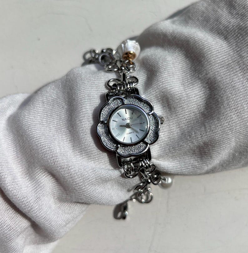 May include: A silver-toned quartz watch with a flower-shaped face and a linked bracelet. The watch face is white with black numerals and hands. The brand name "NAIDU" is printed on the watch face. The bracelet has decorative charms.
