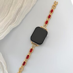 May include: Gold-toned smartwatch band with red oval-shaped accents. The band is connected to a black screen smartwatch. The band has a decorative design and is made of metal. The watch is on a white surface.
