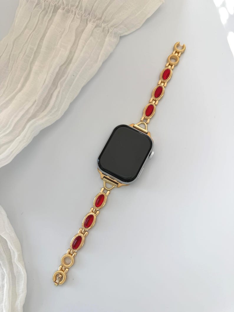 May include: Gold-toned smartwatch band with red oval-shaped accents. The band is connected to a black screen smartwatch. The band has a decorative design and is made of metal. The watch is on a white surface.