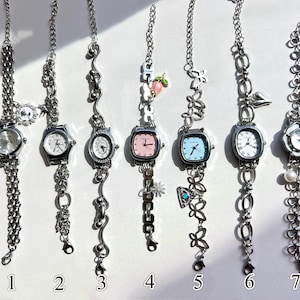 Może przedstawiać: Siedem srebrnych zegarków na łańcuszku z różnymi kształtami tarczy i ozdobnymi zawieszkami. Każdy zegarek ma unikalny design, w tym okrągłe, kwadratowe i owalne tarcze. Niektóre zegarki mają zawieszki, takie jak brzoskwinia, motyle i serca. Zegarki są ponumerowane od 1 do 7.