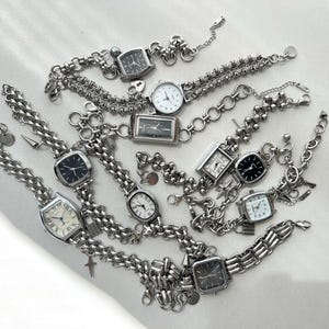 May include: A collection of silver-toned wristwatches with black and white faces, connected by chains and charms. The watches have different styles and sizes, and some have decorative elements like beads and charms.