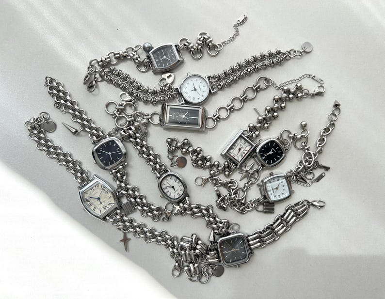 May include: A collection of silver-toned wristwatches with black and white faces, connected by chains and charms. The watches have different styles and sizes, and some have decorative elements like beads and charms.