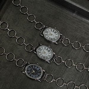 May include: Three silver watches with Roman numeral faces attached to a silver chain with large, round links.