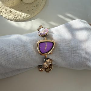 May include: A gold wristwatch with a purple face and decorative charms. The watch has an unusual, asymmetrical shape and is decorated with floral and circular charms. The watch is displayed on a white surface.