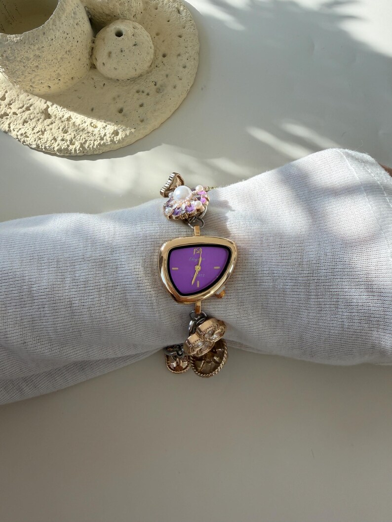 May include: A gold wristwatch with a purple face and decorative charms. The watch has an unusual, asymmetrical shape and is decorated with floral and circular charms. The watch is displayed on a white surface.