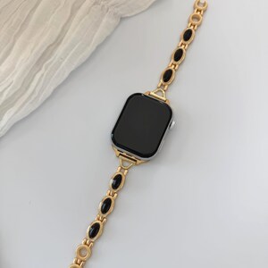 May include: A gold-toned watch band with black oval accents is attached to a smartwatch with a black screen. The band is made of a series of decorative links. The watch and band are set against a white background.
