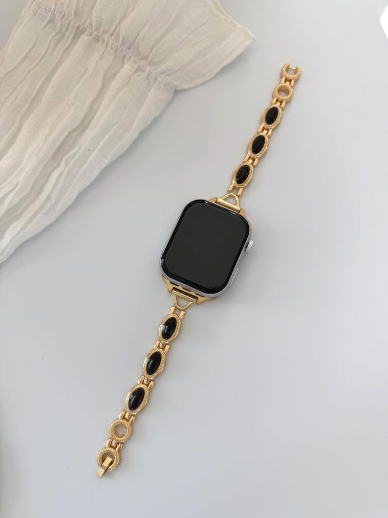 May include: A gold-toned watch band with black oval accents is attached to a smartwatch with a black screen. The band is made of a series of decorative links. The watch and band are set against a white background.