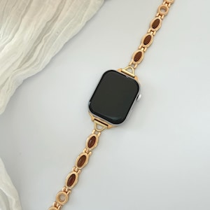 May include: Gold-toned watch band with brown oval accents. The band is connected to a black-screened smartwatch. The band's design features a series of oval-shaped brown stones set within a gold-colored metal frame, creating an elegant look.