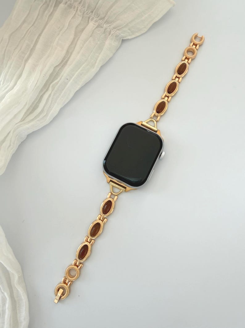 May include: Gold-toned watch band with brown oval accents. The band is connected to a black-screened smartwatch. The band's design features a series of oval-shaped brown stones set within a gold-colored metal frame, creating an elegant look.