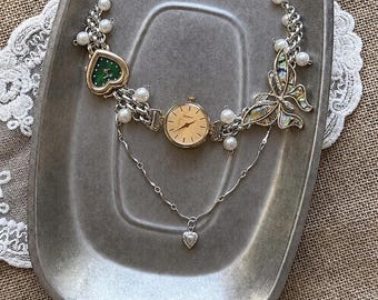 May include: A silver-toned chain necklace featuring a central round watch face, a green heart-shaped watch, and a butterfly charm with iridescent inlay. The design includes white faux pearls, a delicate dangling chain, and a small silver heart charm.