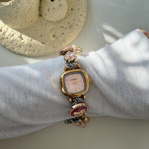 May include: A gold watch with a square pink face and the word "LEAFE" printed on it. The watch band is silver and decorated with charms, including a pink gemstone and a red charm with gold accents. The watch is on white fabric.