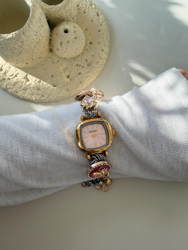 May include: A gold watch with a square pink face and the word "LEAFE" printed on it. The watch band is silver and decorated with charms, including a pink gemstone and a red charm with gold accents. The watch is on white fabric.