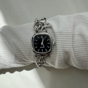 May include: A silver-tone analog wristwatch with a square-shaped case and a black dial featuring the word QUARTZ. The watch has a thick chain-link bracelet adorned with a small, four-pointed star charm attached near the watch case.