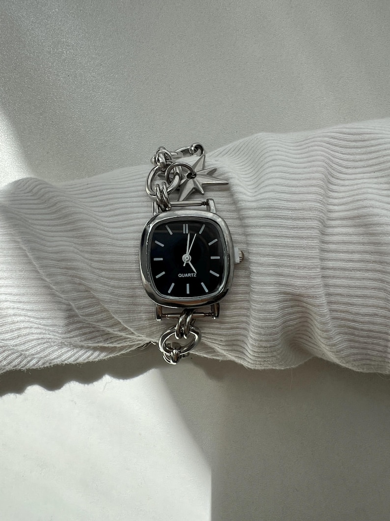 May include: A silver-tone analog wristwatch with a square-shaped case and a black dial featuring the word QUARTZ. The watch has a thick chain-link bracelet adorned with a small, four-pointed star charm attached near the watch case.
