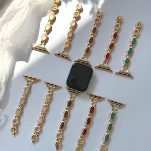 May include: Gold-toned watch bands with various colored oval-shaped gemstones. The bands are arranged around a black smartwatch, showcasing the different color options. The gemstones include shades of beige, brown, red, and green.