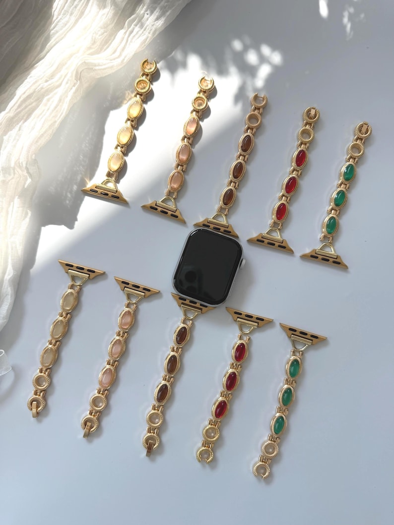 May include: Gold-toned watch bands with various colored oval-shaped gemstones. The bands are arranged around a black smartwatch, showcasing the different color options. The gemstones include shades of beige, brown, red, and green.
