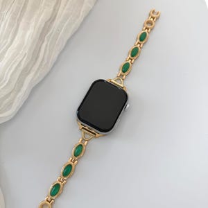 May include: A gold-toned watch band with oval green gemstones. The band is attached to a silver smartwatch with a black screen. The band's design features a series of gold links and green stones, creating a stylish accessory.
