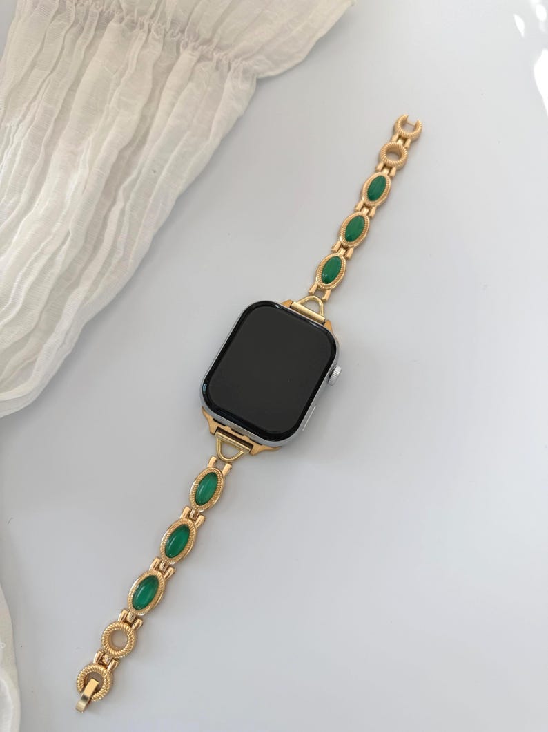 May include: A gold-toned watch band with oval green gemstones. The band is attached to a silver smartwatch with a black screen. The band's design features a series of gold links and green stones, creating a stylish accessory.