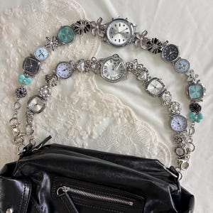 Può includere: Borsa in pelle nera con cinturini per orologi argentati. I cinturini dell'orologio sono adornati con vari quadranti, elementi floreali e perline decorative. La borsa ha una tasca con cerniera e fibbie.