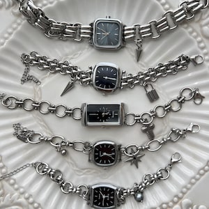 May include: Five silver-toned wristwatches with black or dark gray faces and metal link chains. Each watch features unique charms including spikes, a padlock, a cross, and a star. The watches have analog displays with silver hands and markers.