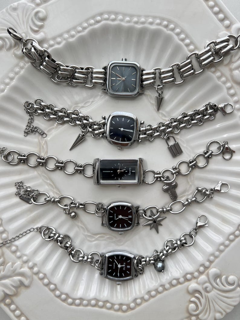 May include: Five silver-toned wristwatches with black or dark gray faces and metal link chains. Each watch features unique charms including spikes, a padlock, a cross, and a star. The watches have analog displays with silver hands and markers.