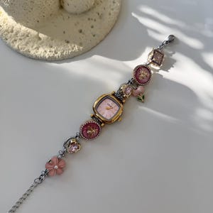 May include: A gold-toned watch with a pink face and decorative charms. The watch band features pink flower and gem accents, with a silver chain. The watch face has the word "LEAF" printed on it.