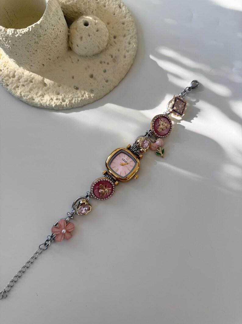 May include: A gold-toned watch with a pink face and decorative charms. The watch band features pink flower and gem accents, with a silver chain. The watch face has the word "LEAF" printed on it.
