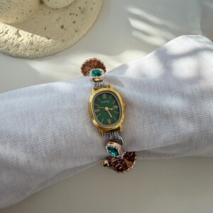 May include: A gold and silver wristwatch with a green face and Roman numeral hour markers. The watch has decorative accents with green gemstones and a textured, leaf-like design. The watch is on a white background.
