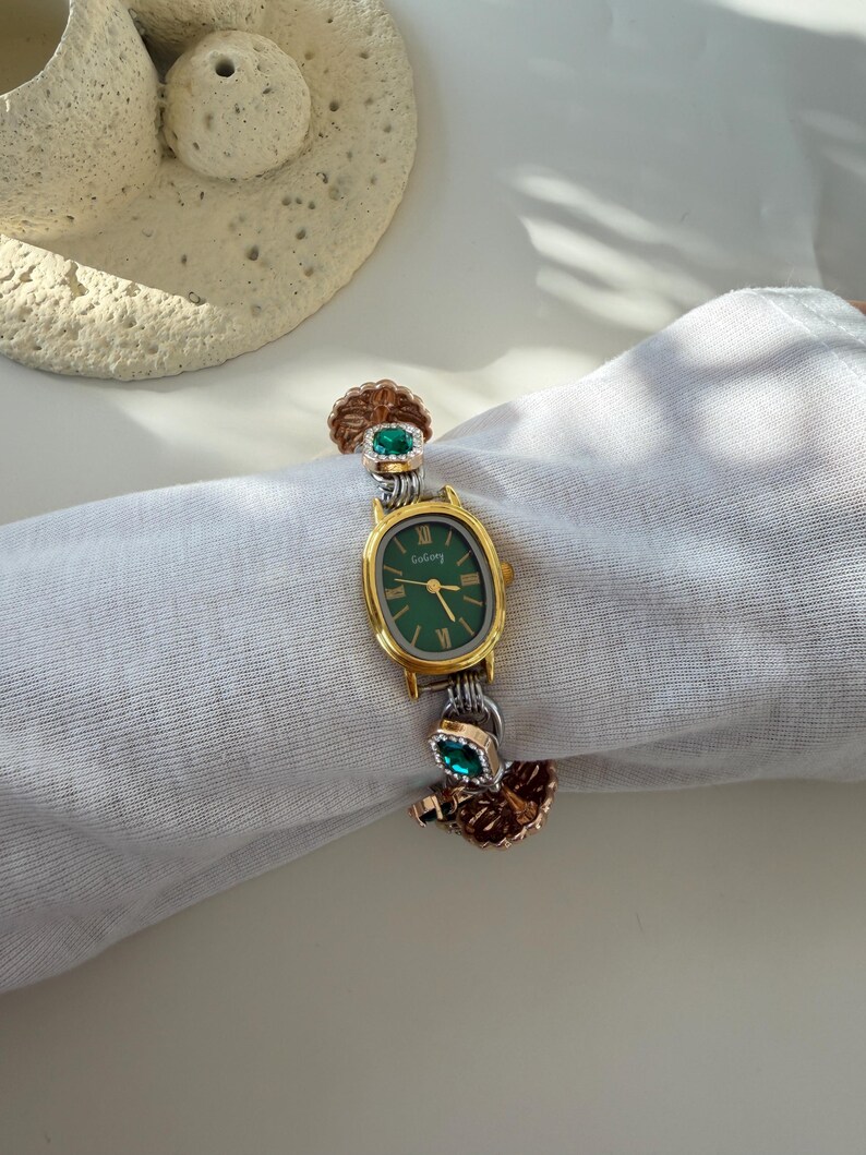 May include: A gold and silver wristwatch with a green face and Roman numeral hour markers. The watch has decorative accents with green gemstones and a textured, leaf-like design. The watch is on a white background.