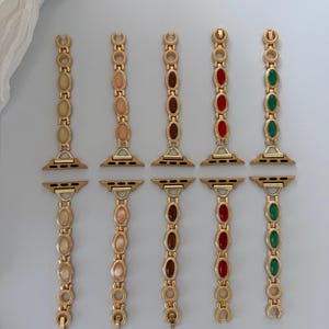 May include: Gold-toned watch bands with various colored oval-shaped gemstones. The bands feature gemstones in shades of beige, pink, brown, red, and green. The bands are designed to attach to a smartwatch.