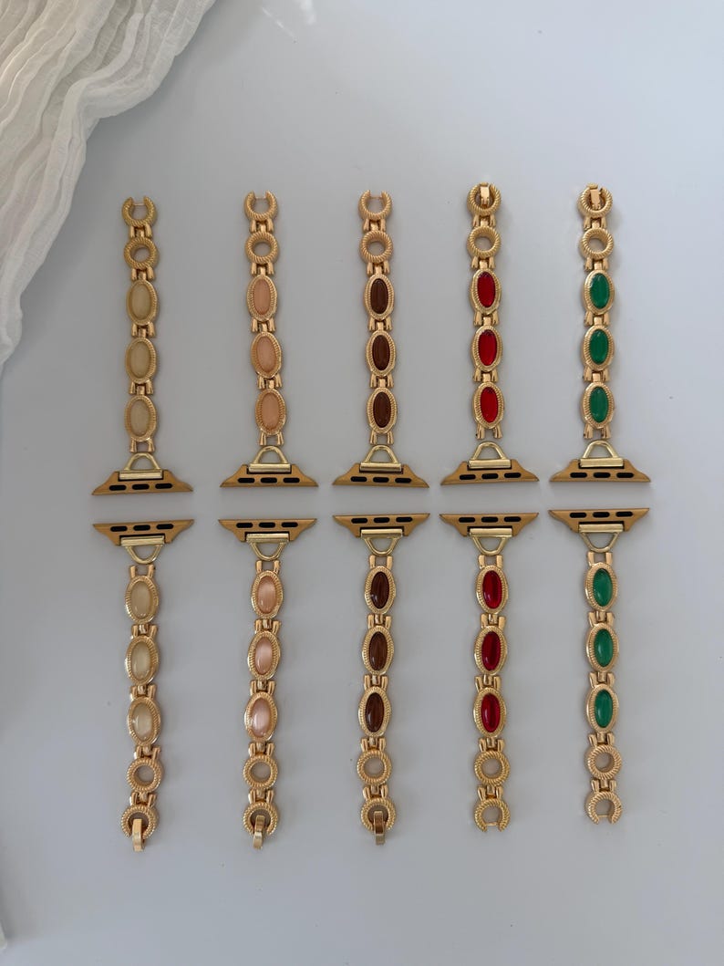 May include: Gold-toned watch bands with various colored oval-shaped gemstones. The bands feature gemstones in shades of beige, pink, brown, red, and green. The bands are designed to attach to a smartwatch.