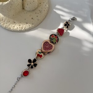 May include: A silver bracelet watch featuring a heart-shaped watch face and decorative charms. Charms include a black flower, a red gem, and heart-shaped designs with cherries and strawberries. The watch has a chain extension.