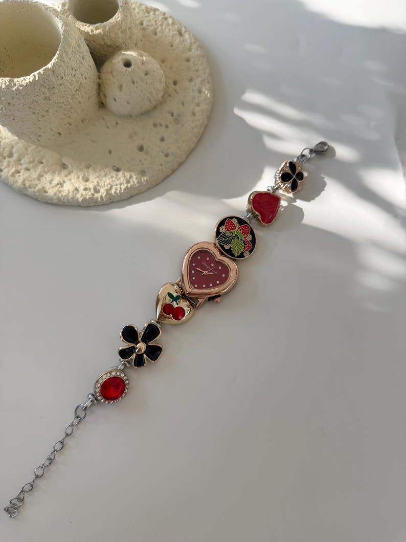 May include: A silver bracelet watch featuring a heart-shaped watch face and decorative charms. Charms include a black flower, a red gem, and heart-shaped designs with cherries and strawberries. The watch has a chain extension.