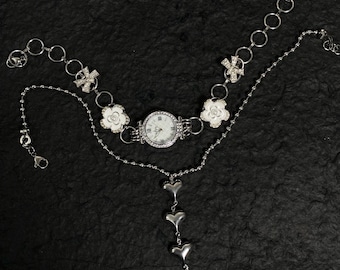 May include: A silver watch with a white face and a silver chain with flower and bow charms. The chain is attached to a silver pendant with three heart charms and a pearl.